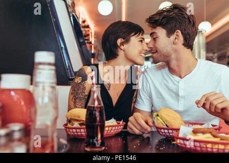 Happy couple at a restaurant holding a french fry in their teeth looking at each other. Man and woman sitting in a diner with food on the table. Stock Photo