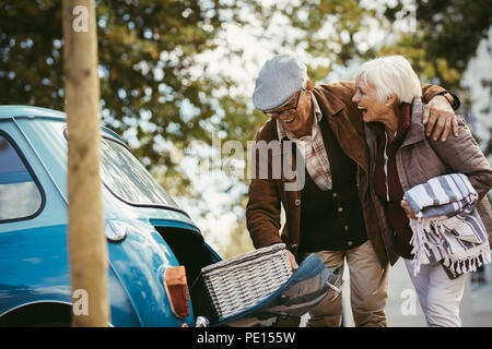Senior couple taking stuff out from the car, preparing for picnic. Laughing old man and woman taking out picnic basket and blanket out of their car tr Stock Photo
