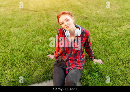 A beautiful red-haired student with freckles is dressed in a red checkered shirt with headphones sitting on the lawn in between the cheba. Student lei Stock Photo