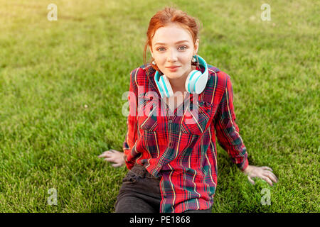 A beautiful red-haired student with freckles is dressed in a red checkered shirt with headphones sitting on the lawn in between the cheba. Student lei Stock Photo