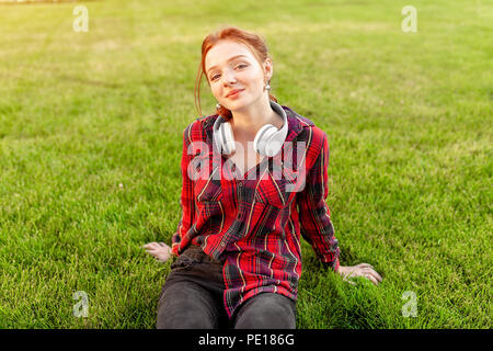 A beautiful red-haired student with freckles is dressed in a red checkered shirt with headphones sitting on the lawn in between the cheba. Student lei Stock Photo