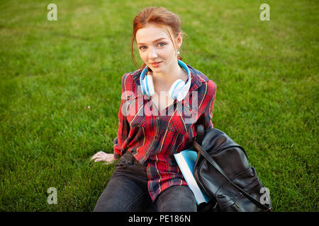 A beautiful red-haired student with freckles is dressed in a red checkered shirt with headphones sitting on the lawn in between the cheba. Student lei Stock Photo