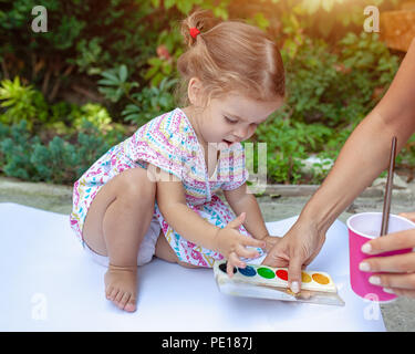 A little girl with curly hair sits on the floor and paints brightly colored paints on a large white sheet of paper. Summer vacation. Leisure for kids. Stock Photo