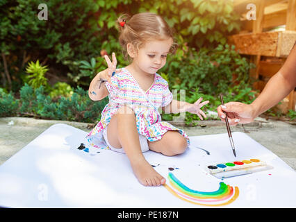 A little girl with curly hair sits on the floor and paints brightly colored paints on a large white sheet of paper. Summer vacation. Leisure for kids. Stock Photo