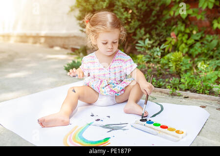 A little girl with curly hair sits on the floor and paints brightly colored paints on a large white sheet of paper. Summer vacation. Leisure for kids. Stock Photo