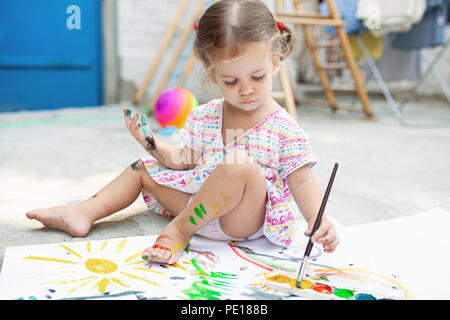 A little girl with curly hair sits on the floor and paints brightly colored paints on a large white sheet of paper. Summer vacation. Leisure for kids. Stock Photo