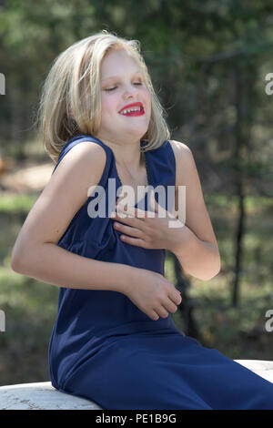 Pretty, young blonde preteen, fashion shoot. Sitting on rock, outside, 3/4 profile, blue dress, laughing, model released. Stock Photo