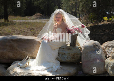 Fantasy, 8-9 year attractie blonde, wearing aunt's wedding dress & veil. Sitting outdoors on large rock beside vase of red roses. Stock Photo