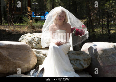 Fantasy, 8-9 year attractie blonde, wearing aunt's wedding dress & veil. Sitting outdoors on large rock beside vase of red roses. Stock Photo