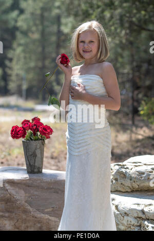 Fantasy, 8-9 year attractie blonde, wearing aunt's wedding dress . Standing outdoors, holding a red rose, beside vase of red roses. Smiling at camera Stock Photo