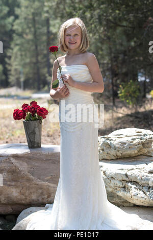 Fantasy, 8-9 year attractie blonde, wearing aunt's wedding dress . Standing outdoors, holding a red rose, beside vase of red roses. Smiling at camera Stock Photo