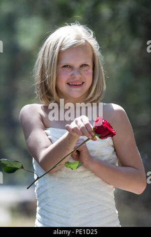 Fantasy, 8-9 year attractie blonde, wearing aunt's wedding dress . Standing outdoors, holding a red rose,  Smiling at camera. Stock Photo