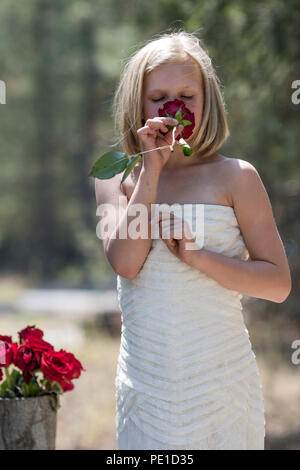 Fantasy, 8-9 year attractie blonde, wearing aunt's wedding dress . Standing outdoors, holding a red rose, beside vase of red roses. Smiling at camera Stock Photo