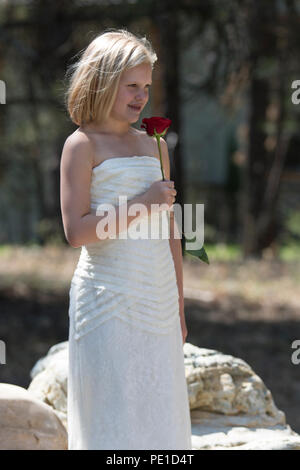 Fantasy, 8-9 year attractie blonde, wearing aunt's wedding dress . Standing outdoors, holding a red rose, #/4 profile looking ca,era right. Stock Photo