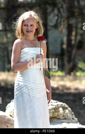 Fantasy, 8-9 year attractie blonde, wearing aunt's wedding dress . Standing outdoors, holding a red rose, Smiling at camera. Stock Photo