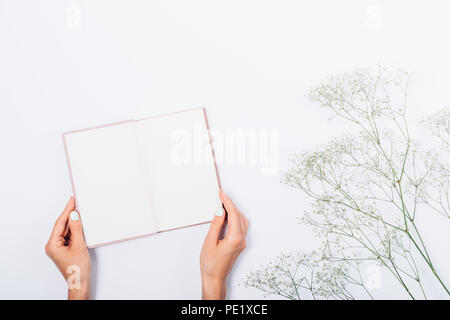 Woman's hands holding open book next to flowers branch on white background, top view. Girl showing blank pages of notebook, flat lay. Stock Photo
