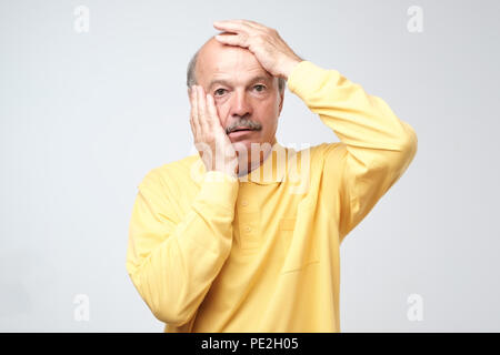 Senior hispanic man in yellow tshirt pulling skin on face with hands. He is scared or tired of stress. I am a little bit crazy concept Stock Photo