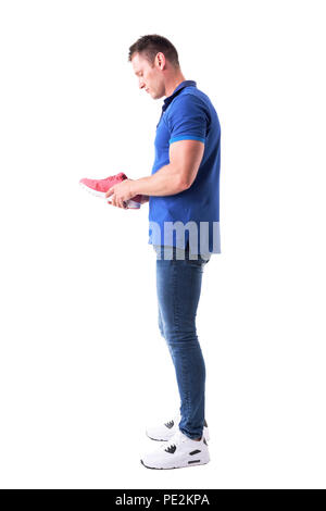 Side view of young adult shopper man holding and examining sneakers before purchase. Full body isolated on white background. Stock Photo