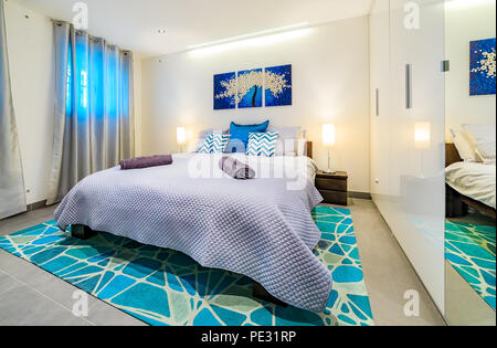 Large bright modern bedroom with a king size bed, decorated in green and blue tones with pillows, wall art painting, rug and welcoming towels Stock Photo
