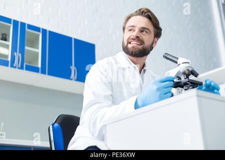 At work. Positive nice male scientist sitting at the table and looking aside while working in the biological lab Stock Photo
