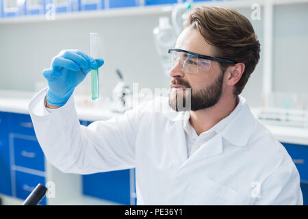 Chemical experiment. Nice positive serious man wearing protective glasses and holding a test tube while doing a chemical experiment Stock Photo