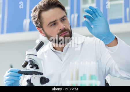 Innovative treatment. Smart handsome nice scientist sitting in front of the microscope and looking at the pill while developing new medicine Stock Photo