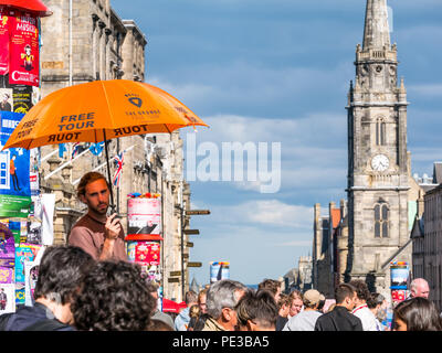 Man advertising free orange tour under umbrella with Tron Kirk spire and clock tower, Royal Mile, Edinburgh, Scotland, UK during Fringe festival Stock Photo