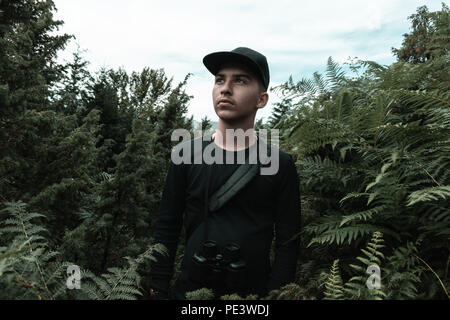 portrait of young fashionable man with binoculars in green bushes and fern plants Stock Photo