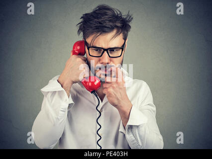 Young frustrated man in glasses having call on telephone and biting nail looking nervous while speaking on gray background Stock Photo