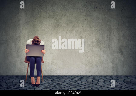 Anonymous woman sitting on chair with laptop on knees and looking desperate in crisis against gray wall background Stock Photo