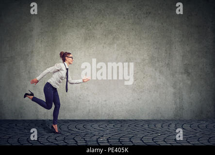 Side view of modern formal business woman running forward under time pressure willing to manage professional competition against gray wall Stock Photo