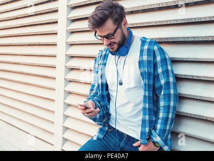 Trendy hipster man in blue shirt and headphones surfing smartphone while leaning on wall on street and smiling Stock Photo