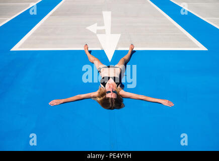 Colour image of a blonde woman in a black bikini lying spreadeagled on her back on blue asphalt, with a white arrow pointing to between her legs Stock Photo