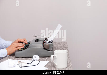 The man typing on an old-fashioned typewriter. On the table are glasses and there is a mug. Neutral background Stock Photo