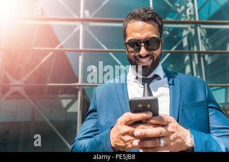 Bearded businessman wears blue suit and sunglasses uses smartphone, typing message before high modern building Stock Photo