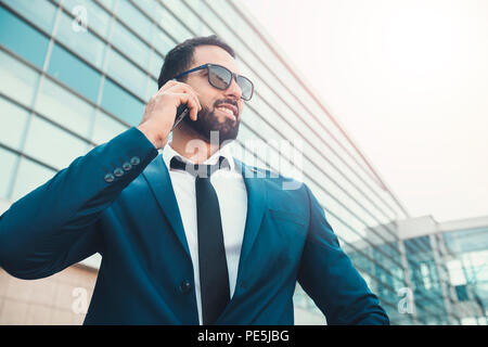 Bearded smiling businessman wears blue suit and sunglasses talking via smartphone before high modern building Stock Photo