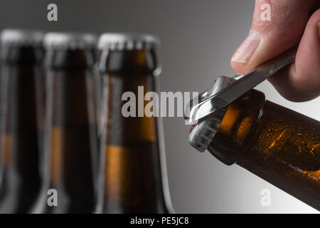 Opening a bottle of beer. Male hand using metal beer opener. Three full bottles against grey wall in blurred background. Stock Photo
