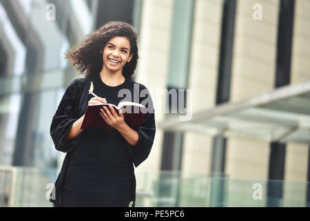 Charming Afro American beautiful happy female, making notes in notepad while walking outdoors on a city street. Education and business concept with sp Stock Photo