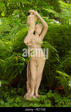 Dione - the divine water goddess, carrying her jar. Stone statue in a cottage garden, between ferns and wild strawberries. Habitat, Belmont, MA, USA Stock Photo