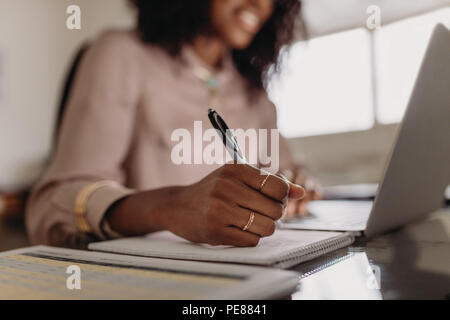 Businesswoman working on laptop computer sitting at home. Close up of hand of a woman holding pen and writing on notepad.