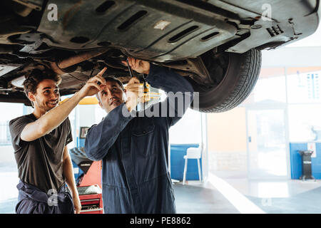 Mechanic fixing the car with coworker pointing and smiling. Two auto repair men working under a lifted vehicle in garage. Stock Photo
