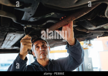 Man tightening a silencer pipe with wrench spanner in auto garage. Mechanic working under a lifted car in repair shop. Stock Photo