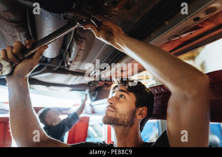 Young man working underneath a lifted car. Mechanic tightening a car part with spanner in automobile service center. Stock Photo