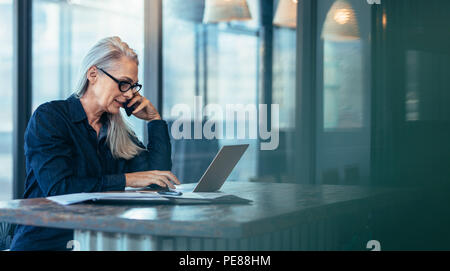 Senior business woman talking on cell phone while working on laptop in office. Mature female using laptop and talking on mobile phone. Stock Photo