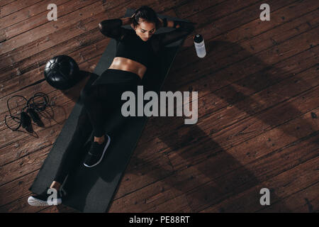 Overhead view of female doing abs workout at the gym. Muscular young woman lying on exercise mat with her hands behind head doing stretches. Stock Photo