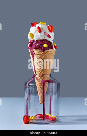 The waffle cone with a cold ice cream stands in glass jar with red and yellow capsules and a lollipop. Jar on a gray background. Pils and lollipops in a jar. Stock Photo