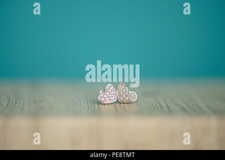 Heart shaped silver pair of earrings with genuine or faux diamonds, displayed on a wooden table with selective focus and blue colorful background Stock Photo