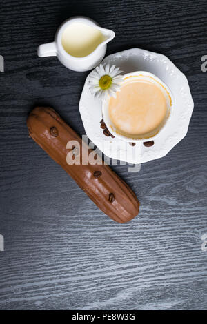 wooden plate with Coffe eclairs with cup of fresh white coffee, pitcher of cream, and coffee beans on Top view. Stock Photo