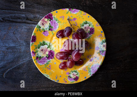 Bunch of washed ripe and juicy red grapes on vine isolated on an ornate yellow plate positioned against a wooden table Stock Photo