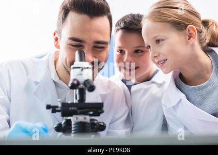 Expecting results. Pleasant joyful merry man using microscope while kids gazeng at him and grinning Stock Photo